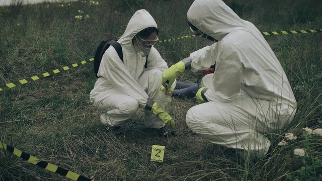 Two people with bacteriological protection equipment picking up evidence into a plastic bag next to corpse outdoors