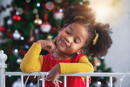 Portrait of little African girl smiling and pointing fingers at her cheeks, against background of Christmas tree, it's X'mas time