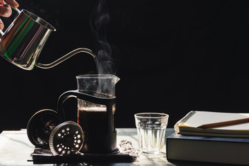 The steam from a pot  to a French press coffee maker on the old wood table and black background with a book,notebook,pencil, Warm drinks make good healthy, Selective focus.