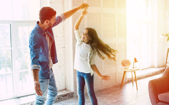 A moment of joy. Tall handsome father is dancing with his cute teenage daughter in their big light living room, laughing and having fun together.