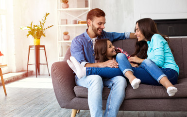 Happy childhood. A beautiful family of father, daughter and mom are sitting on the sofa at home, laughing, hugging and playing with each other.