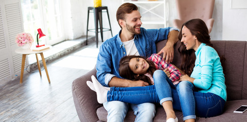 Enjoying the day. Close up photo of young handsome man and his gorgeous wife, who are sitting on the couch in the living-room and hugging their kid.