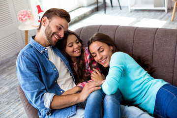 My everything. Close up photo of a beautiful family of mom, daughter and dad, hugging each other on a sofa, smiling kindly and relaxing in the home atmosphere.