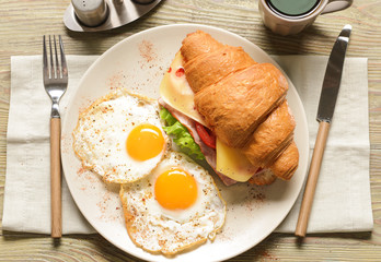 Plate with tasty croissant sandwich and fried eggs on wooden table