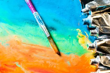 Colored tubes with paintbrush on the painted canvas background. Creative colored wallpaper. Open colored tube on floor. Painted surface. Acrylic Paint dripping from the tube. Painting tools. 