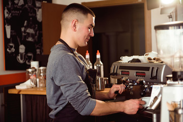 Professional barista holding metal jug warming milk using the coffee machine. Happy young man preparing coffee at counter