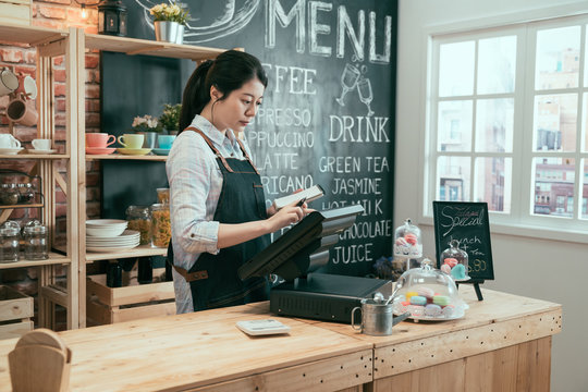 Young Asian Japanese Lady Waitress Using Digital Device For Payment In Coffee Shop Counter. Beautiful Woman Staff Enter Customer Order From Note Into Point Of Sale Terminal Machine In Cafe Store.