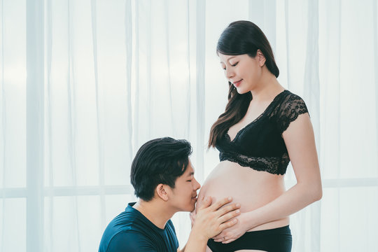 asian chinese man smiling kissing pregnant wife belly with love. young  future parenthood couple. male dad holding abdomen of sexy mom in black  lingerie and pantie by window on happy fathers day.