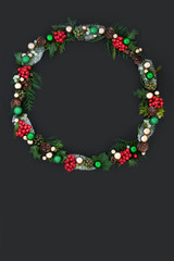 Christmas wreath with winter flora of holly, ivy, mistletoe, cedar and pine cones with gold and green bauble decorations on dark grey background with copy space. 