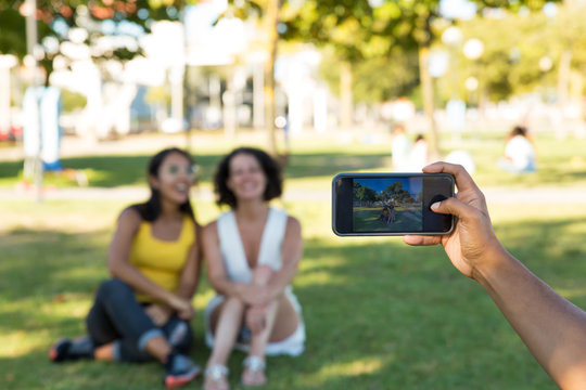 Man photographing female friends in park. Cropped shot of male hand holding smartphone and taking picture of beautiful smiling woman sitting in park. Technology concept