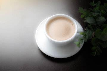 Latte coffee in white cup with saucer, smooth without stripes placed on a black table and small trees beside. Artificial light to replace window light. Blurred, cloud object, give feeling like a dream