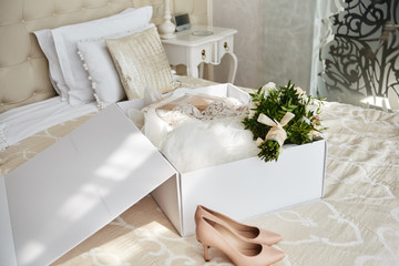 Luxury wedding dress in white box, beige women's shoes and bridal bouquet on bed in hotel room, copy space. Bridal morning preparations. Wedding concept