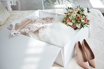 Luxury wedding dress in white box, beige women's shoes and bridal bouquet on bed, copy space....