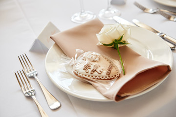 Table setting with blank guest card, white plate with beige serviette, rose, heart and cutlery on table, copy space. Place setting at wedding reception. Table served for wedding banquet in restaurant