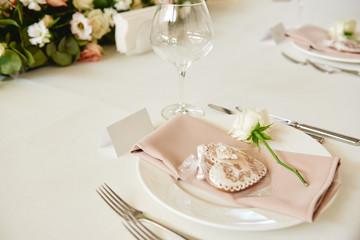Table setting with blank guest card, white plate with beige serviette, rose, heart and cutlery on table, copy space. Place setting at wedding reception. Table served for wedding banquet in restaurant