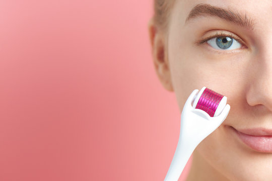 Close-up Female Face With Dermaroller For Mesotherapy Procedures, Skin Care At Home And In Salon. Meso Roller With Microneedles On Pink Background