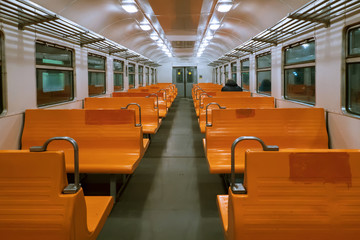 The interior space of a suburban railroad car with orange seats. Evening time, the window is dark. In the background there is a silhouette of one passenger. Background.