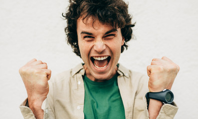 Close-up portrait of successful male with curly hair wearing green t-shirt and beige shirt, screaming with winning expression, fists pumped posing on concrete wall. People and emotion concept