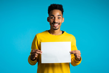 Portrait of young handsome african american man holding white empty paper blank on blue studio background. Copy space. Positive guy with sign