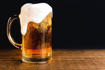 Cold beer with foam in a mug, on a wooden table and a dark background with blank space for a logo or text. Stock Photo mug of cold foamy beer close-up.