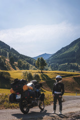Girl in full body protection, turtle armor, knee pads and helmet, motorcycle for traveling with large bags, on a background of green mountains, vertical photo, travel and adventure, autumn day