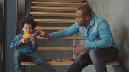 Serious fully engrossed in action adorable mixed race preschool boy playing with toy drill and caring father helping son to use properly work tool on stairs at home, developing child's craft skills.