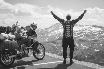 Fototapeta premium A back view motorcycle traveler is happy arrived at his destination. Conquering the top of the mountain, Grossglockner pass, biker dressed in a protective jacket armor black and white. Austria