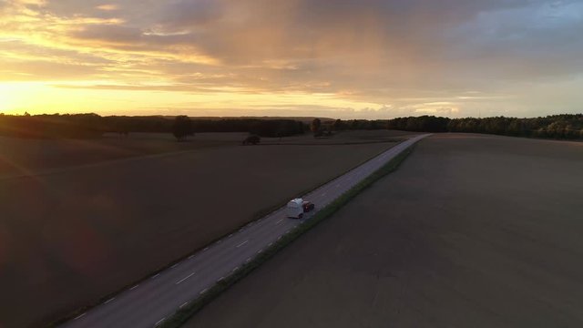 Country road at sunset aerial view. Drone shot flying down over farm fields and road on the countryside, car passing by at dusk