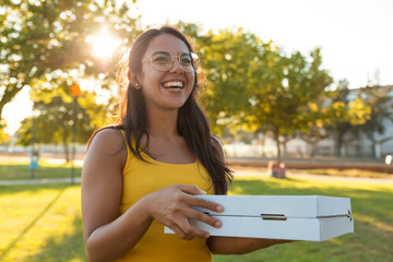 Happy joyful young woman carrying pizza for picnic in park. Beautiful woman standing outdoors, holding pizza boxes, looking away, laughing. Food delivery concept