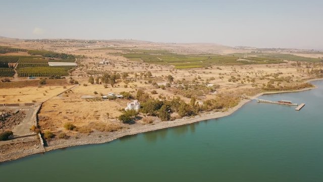 Aeral dolly over Lake Capernaum, shore view, Orthodox Church of the 12 apostles. The coast of Sea of Galilee, top view of the hills, green fields and the road.