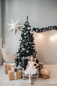 Full length stock photo of lovely little girl in white dress sitting in armchair in front of decorated Christmas tree with presents under it.
