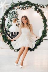 Full length portrait of attractive young adult woman in white dress with bare shoulders and heels sitting on beautiful swings decorated for Christmas. Her daughter decorating Christmas tree in