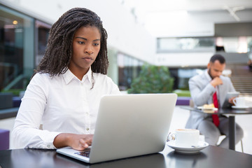 Serious female professional working on laptop while drinking coffee in office lobby. Businessman in formal suit using tablet in background. Workspace in cafe concept