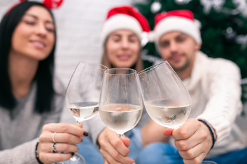 Happy friends celebrating New Year in home interior in Christmas hats sitting near a Christmas tree with glasses of wine. Wine glasses are the focus. Blurred figures of people behind glasses