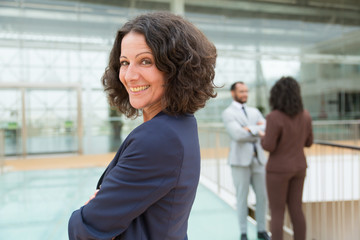 Obraz premium Happy successful business woman posing with arms folded, looking at camera, smiling. Her male and female colleagues standing and talking in background. Confident business leader concept