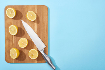 top view of ripe yellow cut lemons on wooden cutting board with knife on blue background with copy...