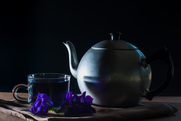 Set of Cup and metal pot of Butterfly pea tea with fresh violet flower on brown tablecloth and wooden table on black background. Healthy beverage for drink. Herbs and medical concept.