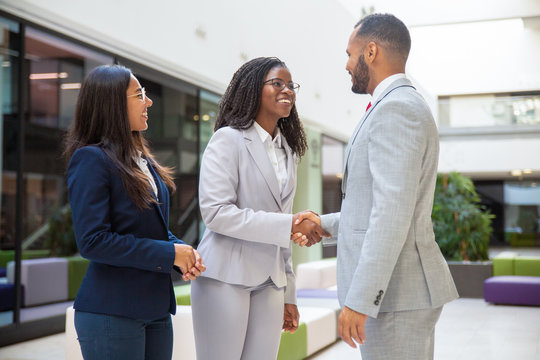 Happy Diverse Business Partners Greeting Each Other. Business Man And Women Standing In Office Hall, Shaking Hands, Talking, Smiling. Partners Meeting Concept