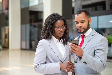 Positive focused colleagues looking at mobile phone screen together. Business man showing smartphone content to female coworker. Mobile internet connection concept