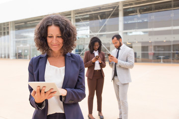Obraz premium Happy satisfied business woman using tablet outside office building. Her male and female colleagues using mobile phones in background. Wireless internet connection concept