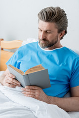 Fototapeta premium handsome patient in medical gown reading book in hospital
