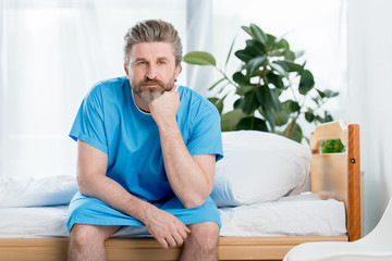 pensive patient in medical gown sitting on bed in hospital