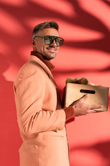 happy man in blazer and sunglasses holding box with sweet peaches on red with shadows