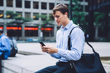 Experienced male director of enterprise sitting outdoors in financial district and using 4g connection for reading news at website, Caucasian businessman in electronic earbuds chatting via app