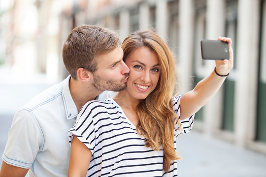 Image of smiling young couple taking selfie photo on cellphone and kissing in the street. She´s looking at the camera