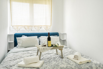 Interior of a bedroom n blue and white colors. Little coffee-table with bottle of wine, glasses and dessert on a bed