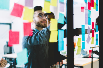 Positive man in optical spectacles with cute smile on face making notes on paper sticks during...