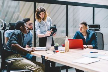 Fototapeta premium Concentrated female manager showing to employees digital information on smartphone, young colleagues reading news in social network while having meeting in office interior sitting together at table