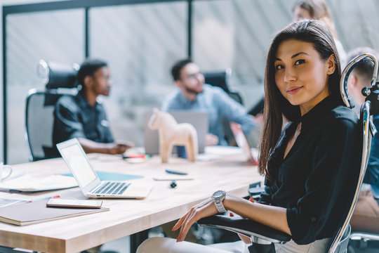 Portrait of Asian prosperous employee sitting front laptop computer at meeting table with male and female colleagues collaborating on blurred background, female office worker satisfied with her job