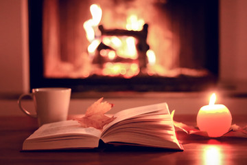 Cozy autumn evening at fireplace at home. Close up photo of open book with fall leaf, cup of tea and candle with fire at background.